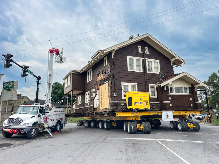 Utility company workers lift powerpoints while Wolfe drives house under them