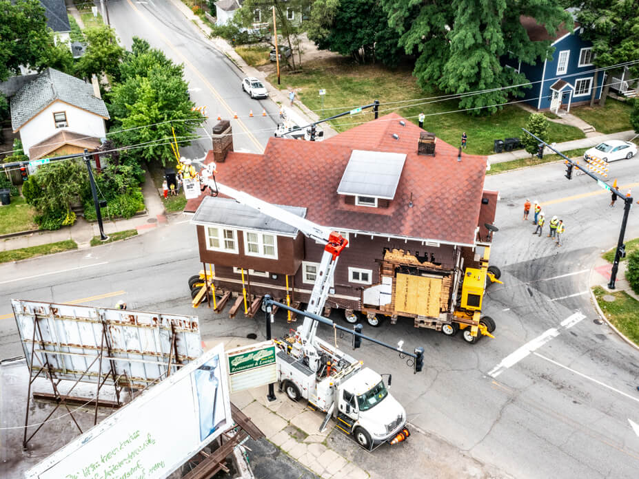 Aerial view of Wolfe steering a house around a corner while workers lift power lines over it