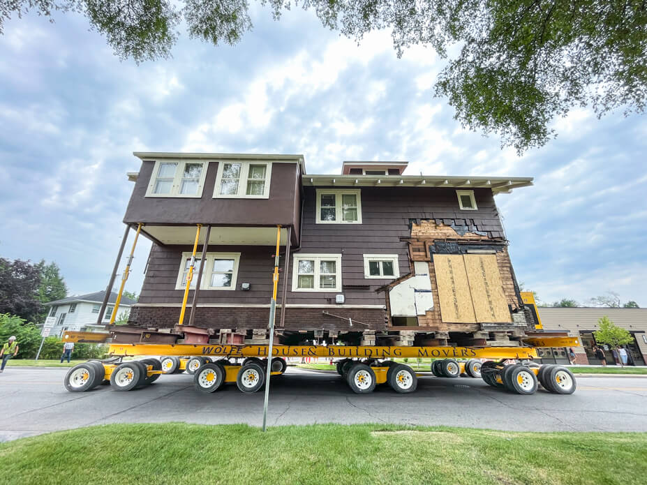 A brown Craftsman-style house sits on steel beams and dollies in the street