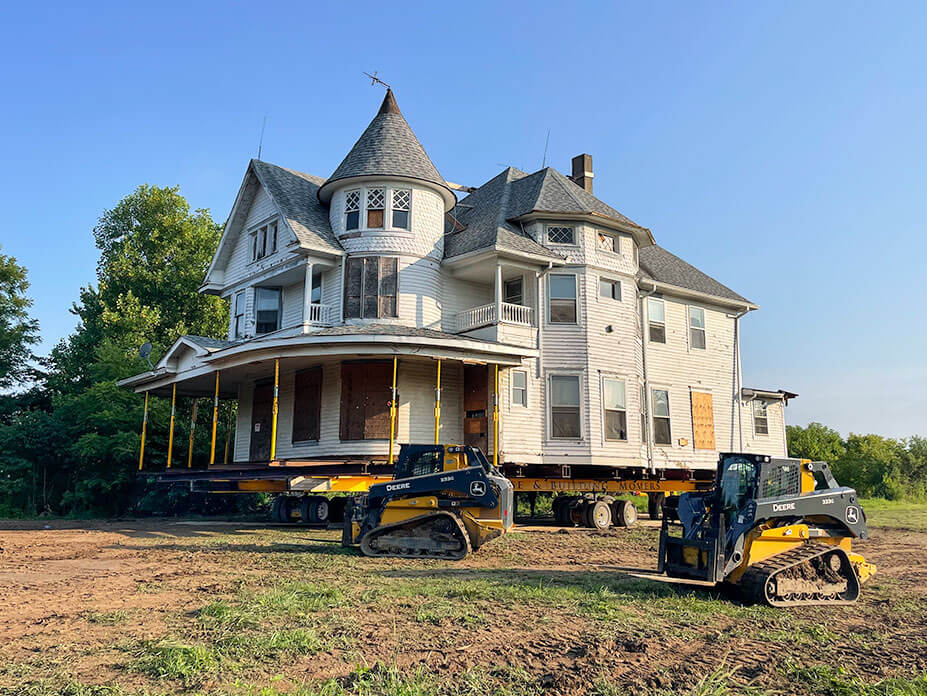 white Queen-Anne style house sit on wheels with two skidsteers in the foreground