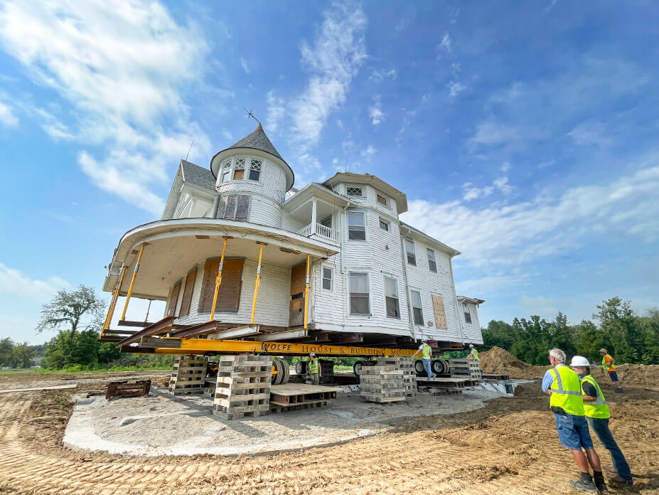 Workers look at a historic Queen-Anne-style house sitting on beams and cribbing over its new foundation