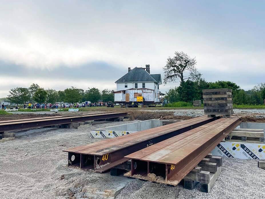 Steel beams lay over house foundation as house drives toward them in background