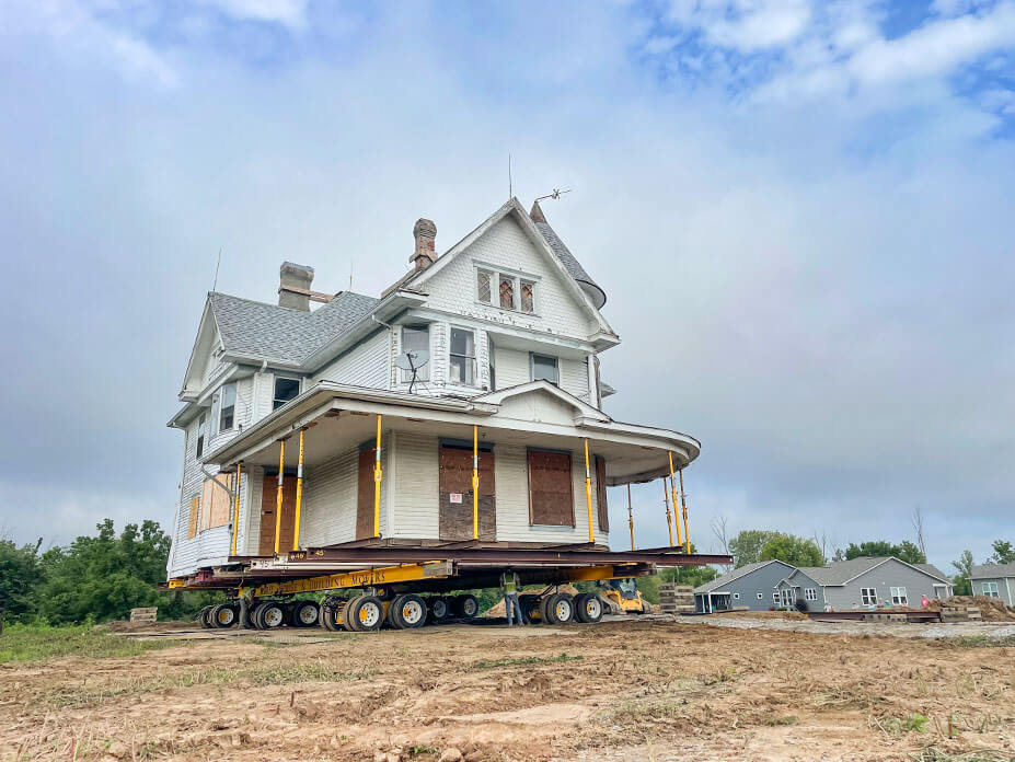 White house on house-moving dollies with wrap-around porch supported by yellow jacking-shoring posts