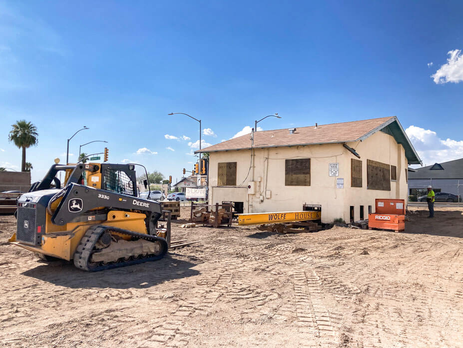 Small cream-colored bungalow with yellow steel beams under it and skid steer in foreground