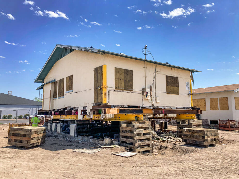 Cream colored bungalow sits on steel beams and cribbing in preparation for relocation