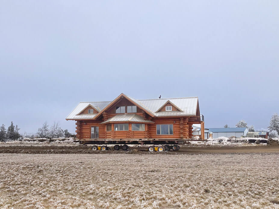 Field in foreground with log cabin on house-moving dollies in background