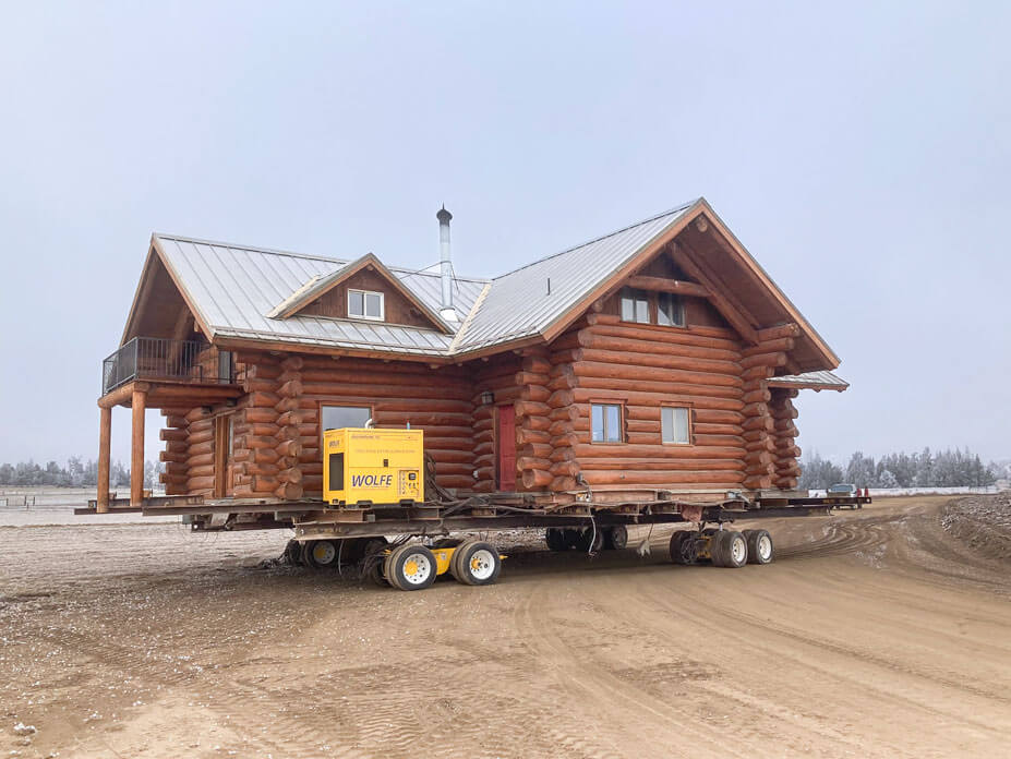 A log cabin with Wolfe power unit and dollies sits in dirt road enroute to new location